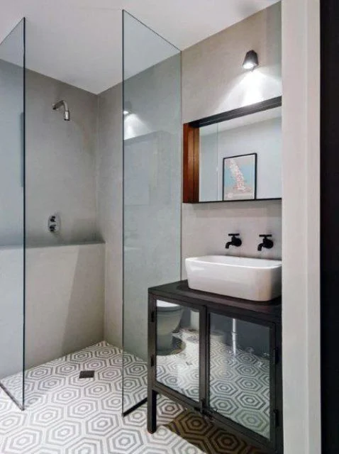 Beton duvarlı ve desenli zemin karolarıyla küçük bir banyo, küçük duş alanı, siyah lavabo ve tezgâh.
