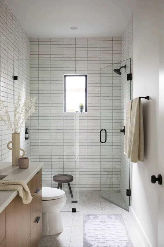 Beyaz ince karolar, duş alanı, lekeli lavabo, siyah armatürler ve küçük pencereye sahip nötr küçük banyo.