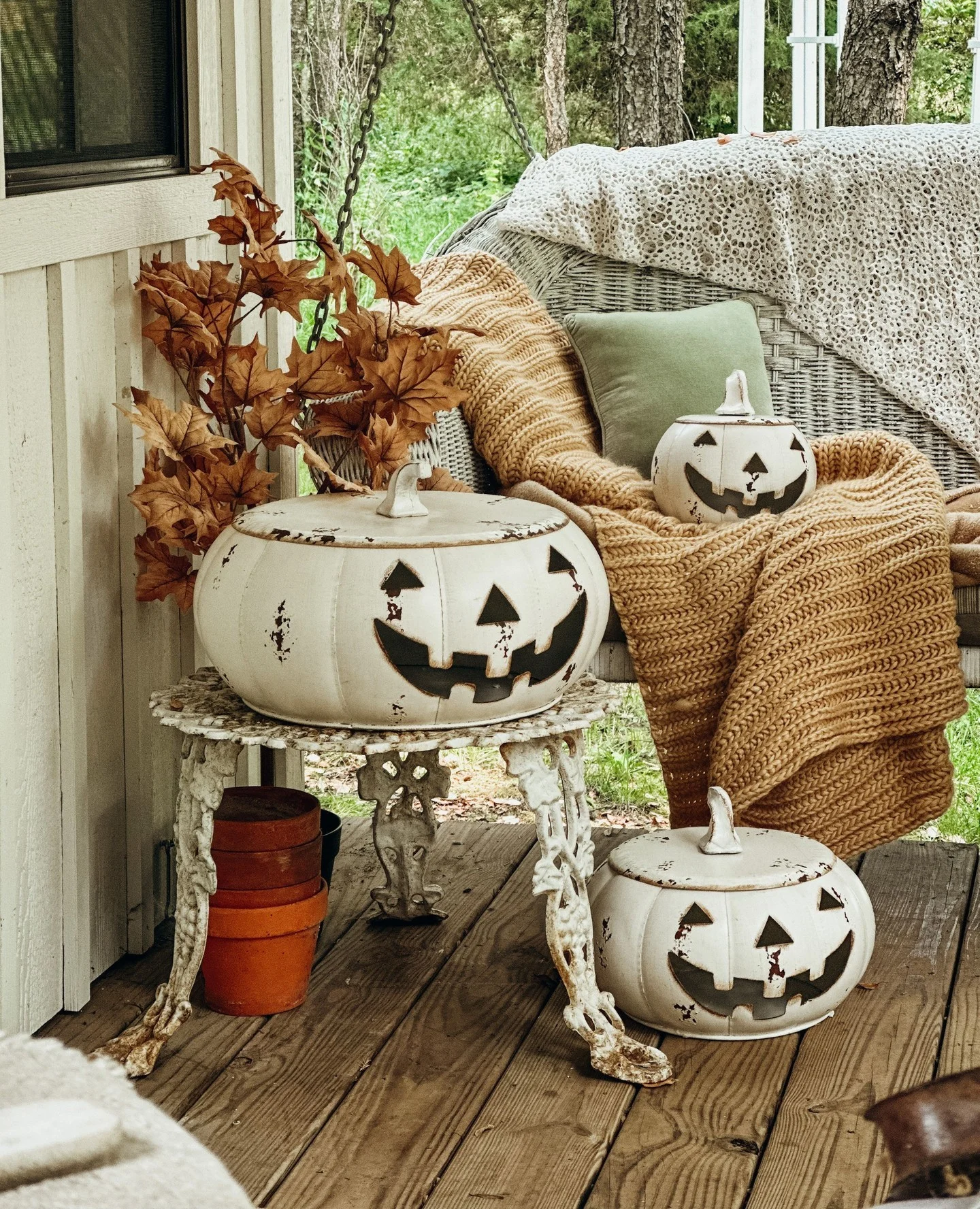 Rahat sonbahar verandası, beyaz jack-o’-lantern kabakları, hardal renkli örgü battaniye ve rustik hasır sandalyede sonbahar yapraklarıyla şirin bir Halloween dekoru.