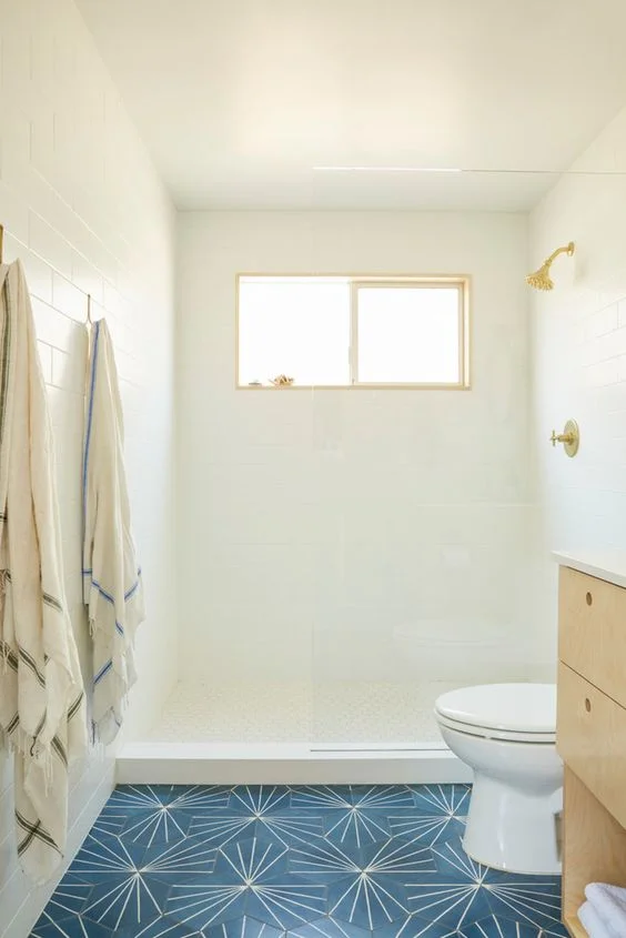 Duvar ve tavanlarda beyaz, zeminde lacivert fayanslar, açık renkli ahşap lavabo ve küçük penceresiyle davetkar banyo.