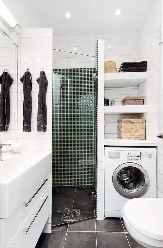 Seramik döşeli duş alanı, çamaşır makinesi, beyaz lavabo ve tezgâh olan çağdaş küçük banyo.