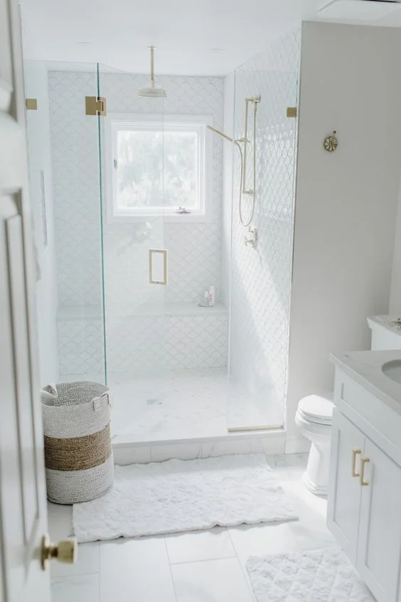 Büyük ve balık pulu desenli beyaz karoların kullanıldığı küçük beyaz banyo, duşta beyaz lavabo, sepet ve altın armatürler.