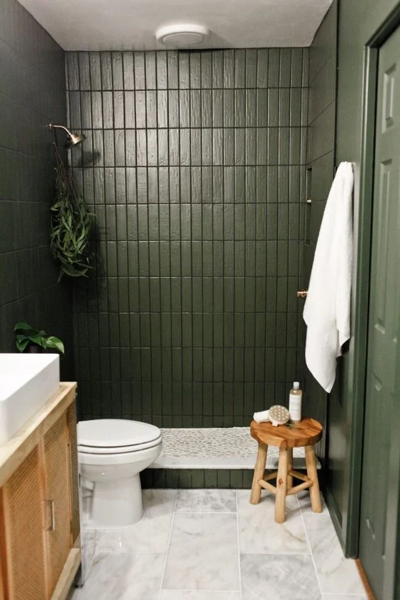 İnce yeşil karolar, kamış lavabo, ahşap tabure, bitkiler ve beyaz havlularla küçük banyo.