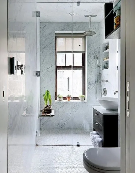Duşta mavi taş karolar, koyu renk lavabo, saksı bitkileri ve gömme raflar bulunan küçük banyo.
