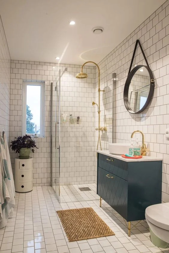 Beyaz fayanslı, küçük ve ferah banyo. Lacivert lavabo, bitkili küçük sehpa ve altın dokunuşlar bulunuyor.
