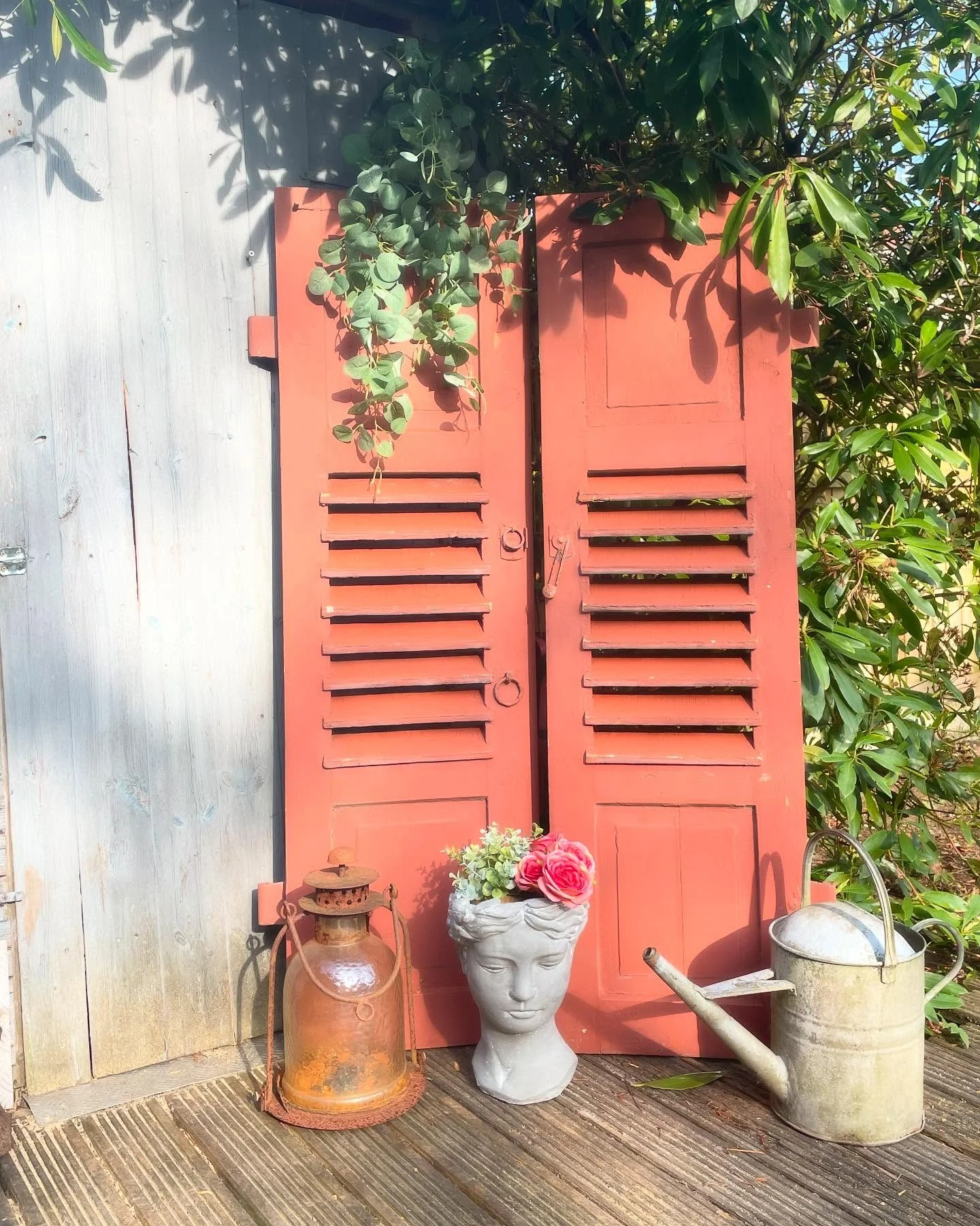Terracotta rengi boyanmış eski panjurlar, sade bir baraka duvarını tarz sahibi bir bahçe köşesine dönüştürüyor. Eskitilmiş fener, güllerle dolu taş saksı ve çinko sulama kabı ile anında kır evi havası yaratılır.