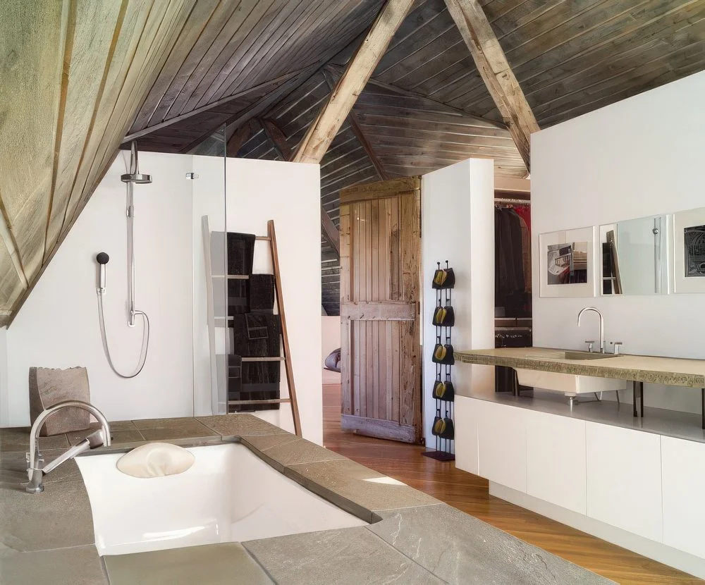 Beyaz duvarlı, kirişli ahşap tavanlı, taş kaplı küvet ve yükseltilmiş taş tezgah olan modern rustik banyo.