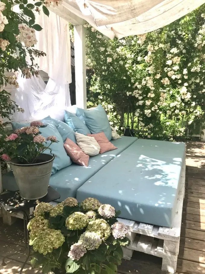 Bu romantik bahçe köşesinde, çiçeklerin tam ortasında mavi döşemeli ve tül örtülü bir daybed yer alıyor.
