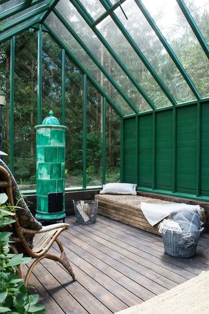 Büyük sera, yeşil kalpli, daybed ve rattan sandalyelerle minimal bir dinlenme alanı. Orman manzarası mekana hava katıyor.