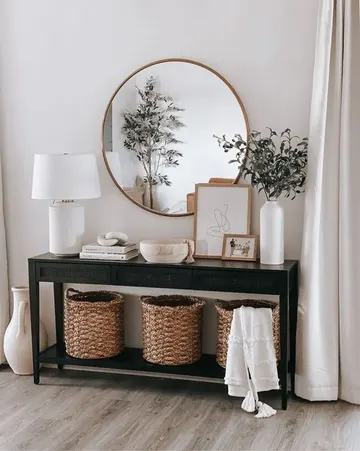 Siyah ahşap konsol, üç depolama sepeti, kitaplar, sanat objesi ve kase, beyaz masa lambası ile yapraklı dallar içeren vazoyla tamamlanmış.