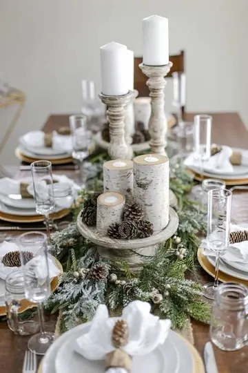 Tüylü çam koşucu, kozalaklar, dal mumluklar ve kozalak peçete halkalarının yer aldığı karla kaplı rustik Noel masası.