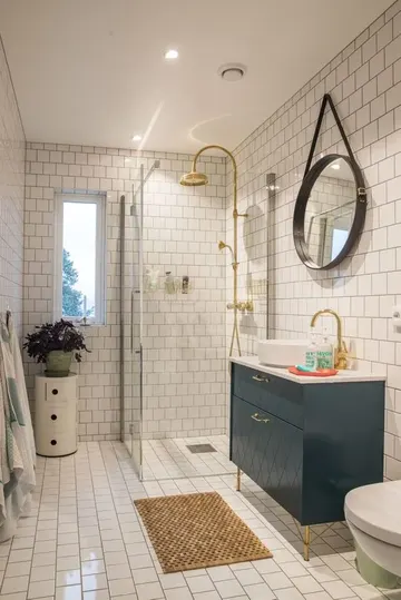 Beyaz fayanslı, küçük ve ferah banyo. Lacivert lavabo, bitkili küçük sehpa ve altın dokunuşlar bulunuyor.