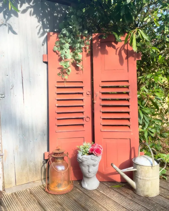 Terracotta rengi boyanmış eski panjurlar, sade bir baraka duvarını tarz sahibi bir bahçe köşesine dönüştürüyor. Eskitilmiş fener, güllerle dolu taş saksı ve çinko sulama kabı ile anında kır evi havası yaratılır.