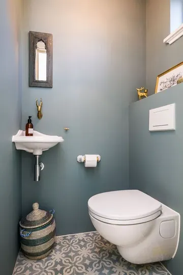 Küçük ve sade bir misafir tuvaleti, küçük duvara monte lavabo, küçük ayna, depolama sepeti ve tuvaletle.