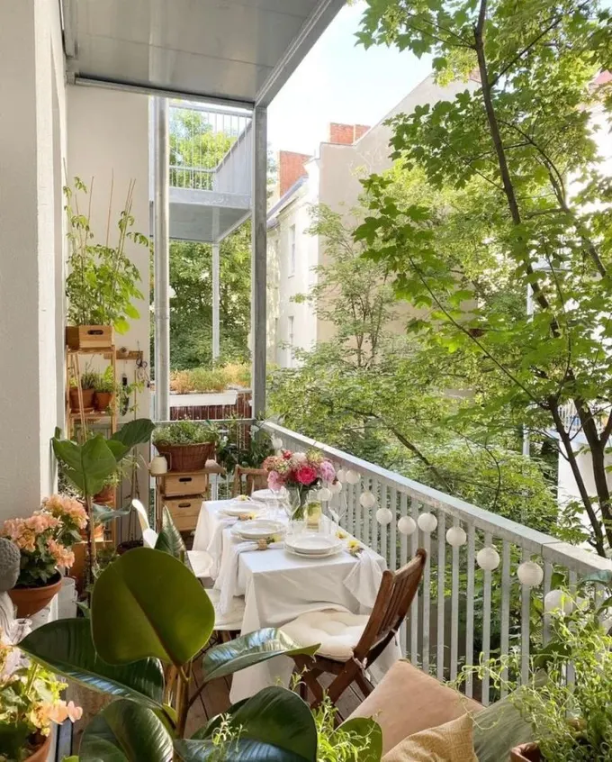 Uzun ve dar balkon, saksı bitkileri, çiçekler ve ışıklarla atmosfer oluşturuyor. Uzun masa katlanabilir sandalyelerle tamamlanmış.
