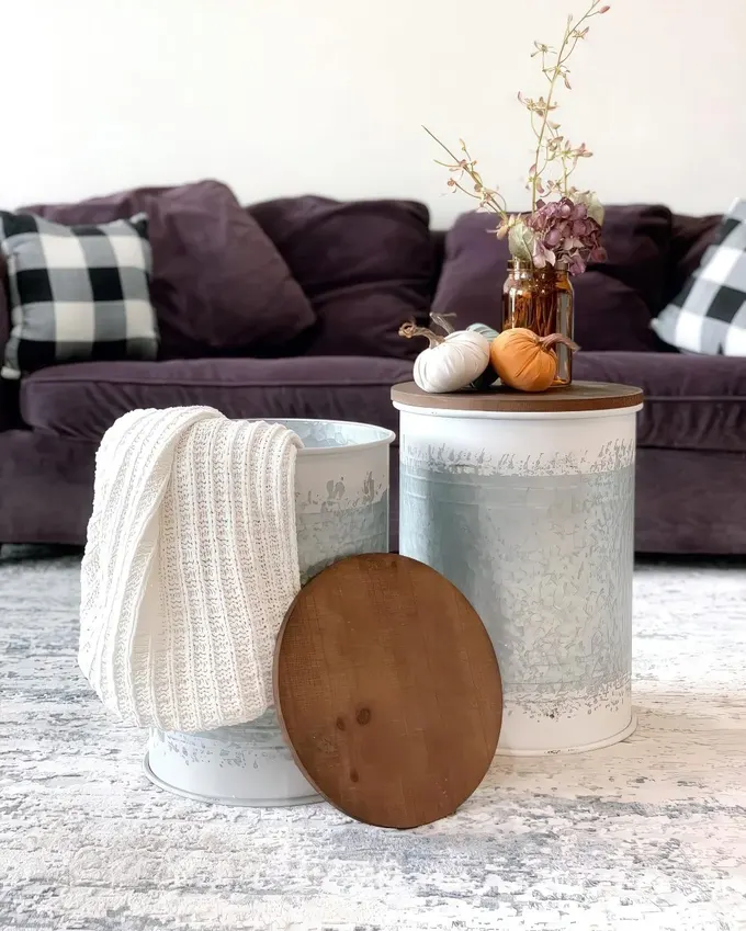 Ahşap kapaklı galvaniz metal variller, şık depolama ve rustik dekor olarak kullanılmış. Beyaz örme battaniye, küçük çiçek aranjmanı ve mini balkabakları mekana çekici bir dokunuş ekliyor.