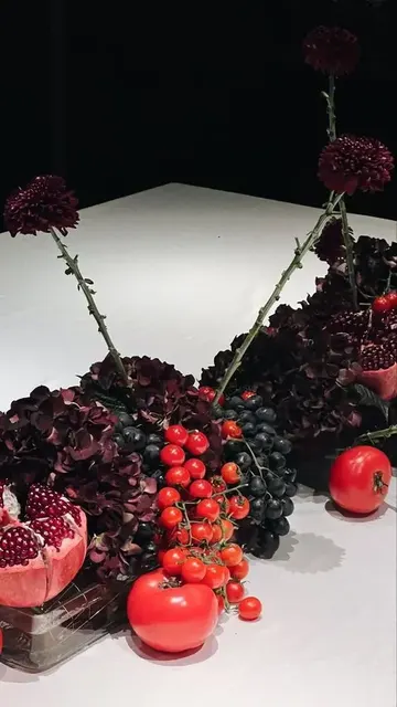 Domatesler, nar, koyu ortancalar, üzüm ve koyu renkli dalyalarla yapılan sonbahar veya Şükran Günü için etkileyici atmosferik bir masa ortası.