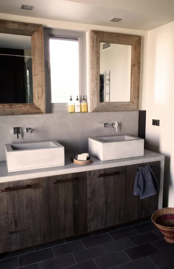 Minimalist betondan duvarlı, yıpranmış ahşap lavabo ünitesi, beton lavaboları ve ahşap çerçeveli aynalarla ahır banyosu.
