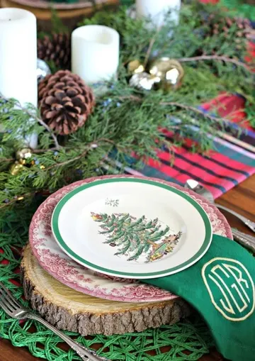 Ahşap dilim tabak altlıkları, desenli tabaklar, çam dalları, kozalaklar, mumlar ve yeşil peçetelerin olduğu geleneksel Noel masası.