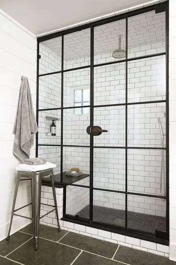 Beyaz metro ve grafit gri karolarla vintage esintili banyo, siyah çerçeveli duş kapıları ve içinde oturma bankı mevcut.