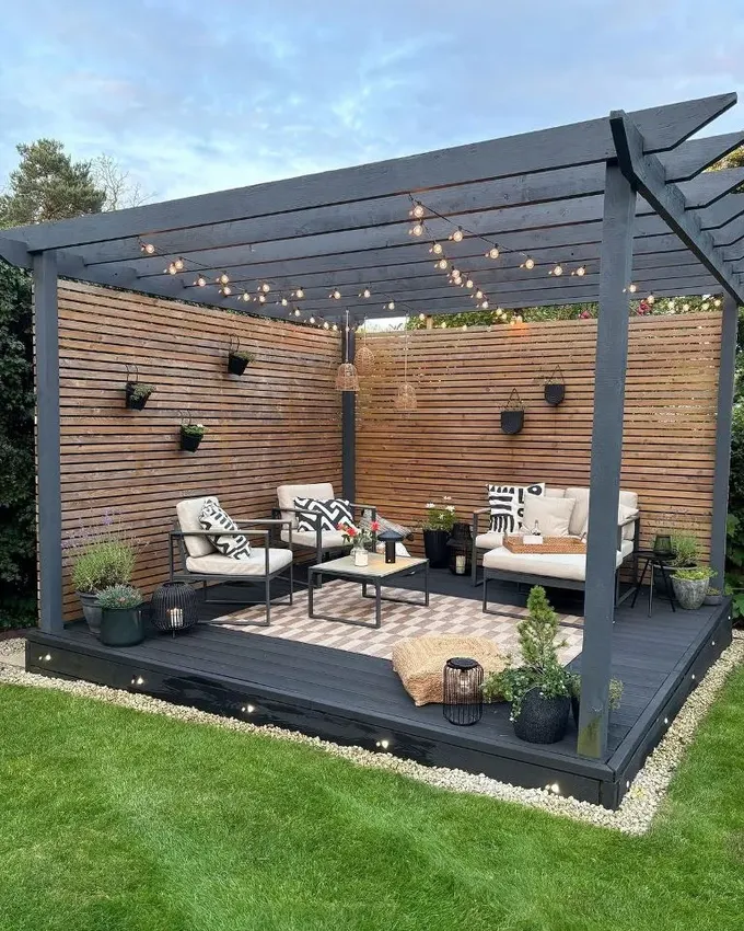 Koyu renk yükseltilmiş güverte, perdeli ve ışıklı pergola ile birleşiyor; modern mobilyalar ve bitkiler hoş bir açık oturma alanı yaratıyor.