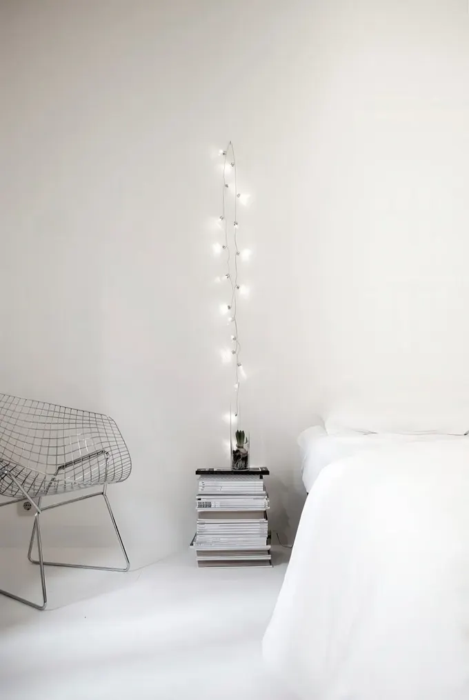 Nötr yatak örtülü alçak yatak, metal sandalye, dergi yığını ve klasik masa lambası yerine üzerine asılan string ışıklarla minimalist İskandinav yatak odası.