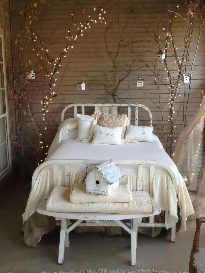 Ahşap panelli taupe renk aksan duvar, beyaz yatak ve bench, dallar ve ağacın üzerindeki ışıklarla parlak bir shabby chic yatak odası.
