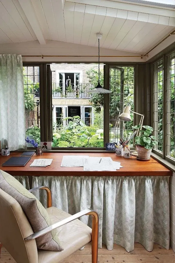 Kır evi tarzındaki bahçe kulübesi, perdesi olan bir masa, rahat bir sandalye ve bahçe manzarasıyla ev ofisi olarak kullanılıyor.