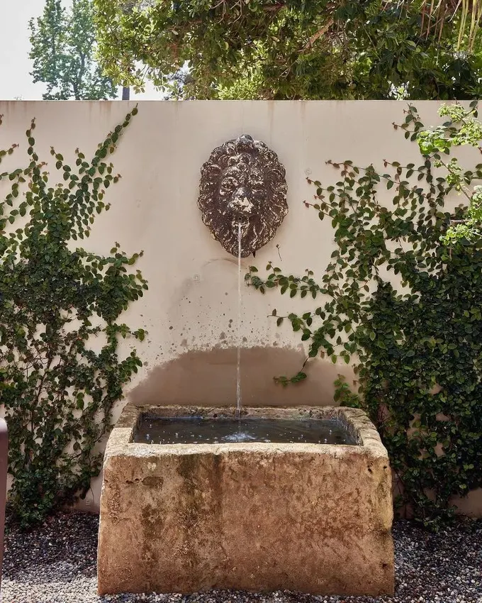 Duvara yerleştirilmiş yüksek kanal, musluğu gizler ve sadece vintage bir madalyon görülür. Yeşil sarmaşıklar çeşme çevresini kaplamıştır.