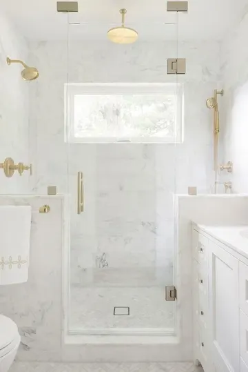 Beyaz mermerli rafine banyo, duşta yarım duvarlar ve altın dokunuşlarla sofistike bir görünüm.