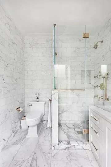 Beyaz mermer fayanslı şık banyo, duş alanında alçak duvar, beyaz lavabo ve altın dokunuşlarla zarafet.