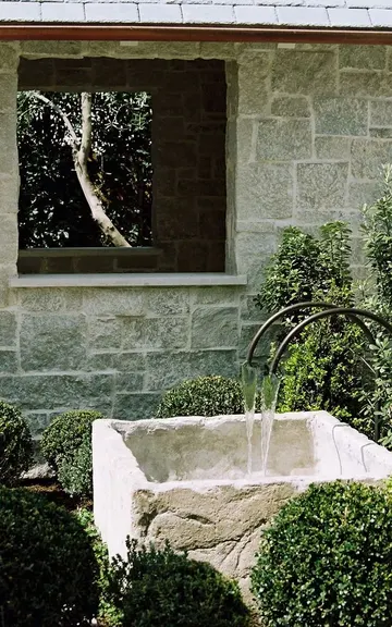 Beyaz çift fıskiye, taşa yumuşaklık katmak için budanmış ağaçlar ve yeşilliklerle çevrili.