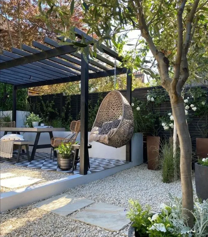 Arka bahçede pergola ve asılı bir yumurta sandalyesi bulunuyor. Uzun ağaçlar hassas ama güneşten koruyor.