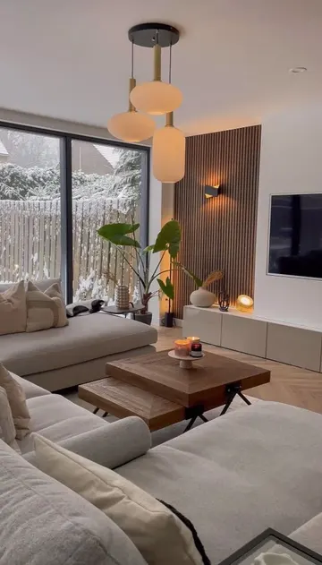 Bu küçük çağdaş oturma odasında nötr köşe koltuk, birkaç masa, TV ve katmanlı aydınlatma var.