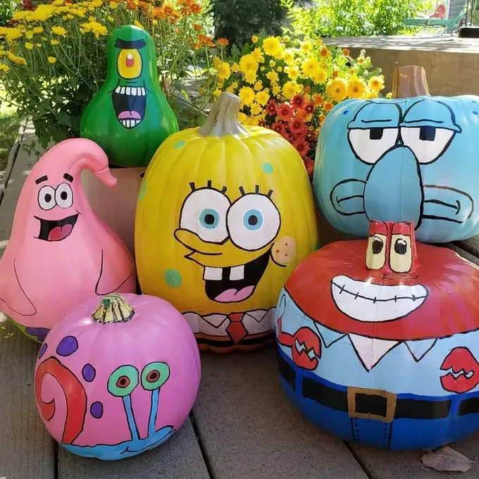 SpongeBob karakterleri Patrick, Squidward, Gary ve Mr. Krabs'ın boyalı renkli kabakları, sarı krizantem çiçekleriyle çevrili; aileler ve çocuklar için eğlenceli kabak süsleme fikri.