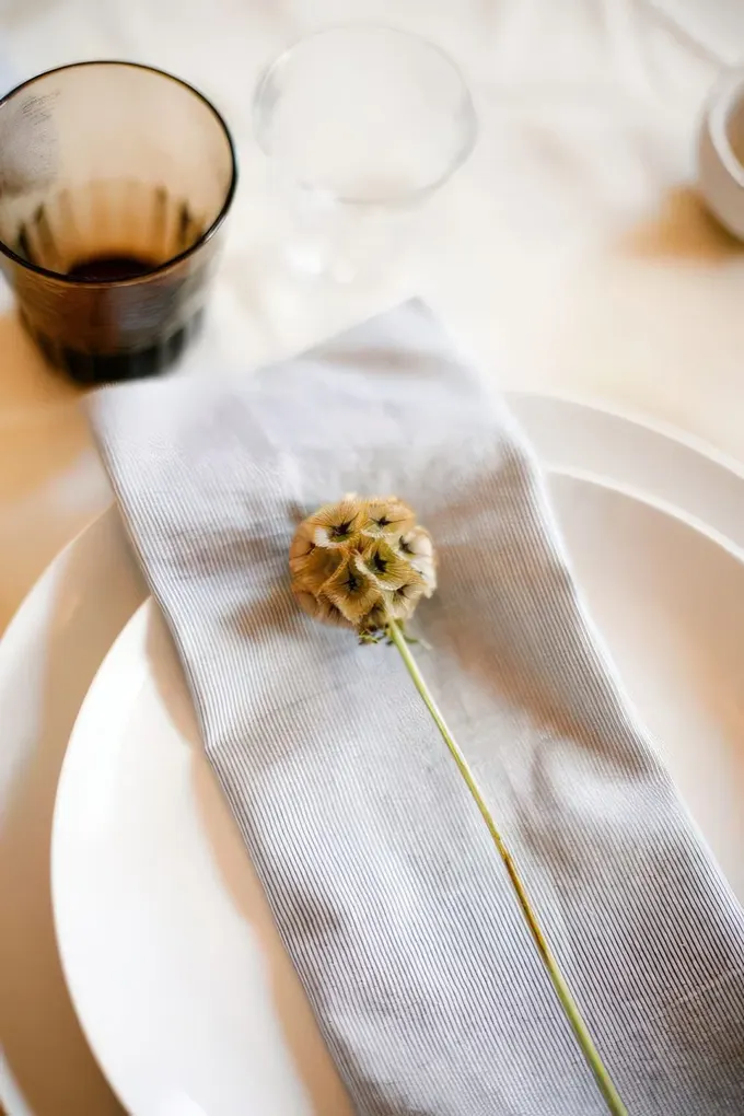 Minimal masa düzeni: parlak beyaz tabaklar, yumuşak gri peçete, tek kurutulmuş çiçek—basit, modern baby shower masası.