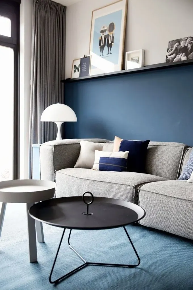 Modern salon, renk bloklu duvar, gri düşük koltuk, sanat rafı ve birkaç masa ile düzenlenmiş.
