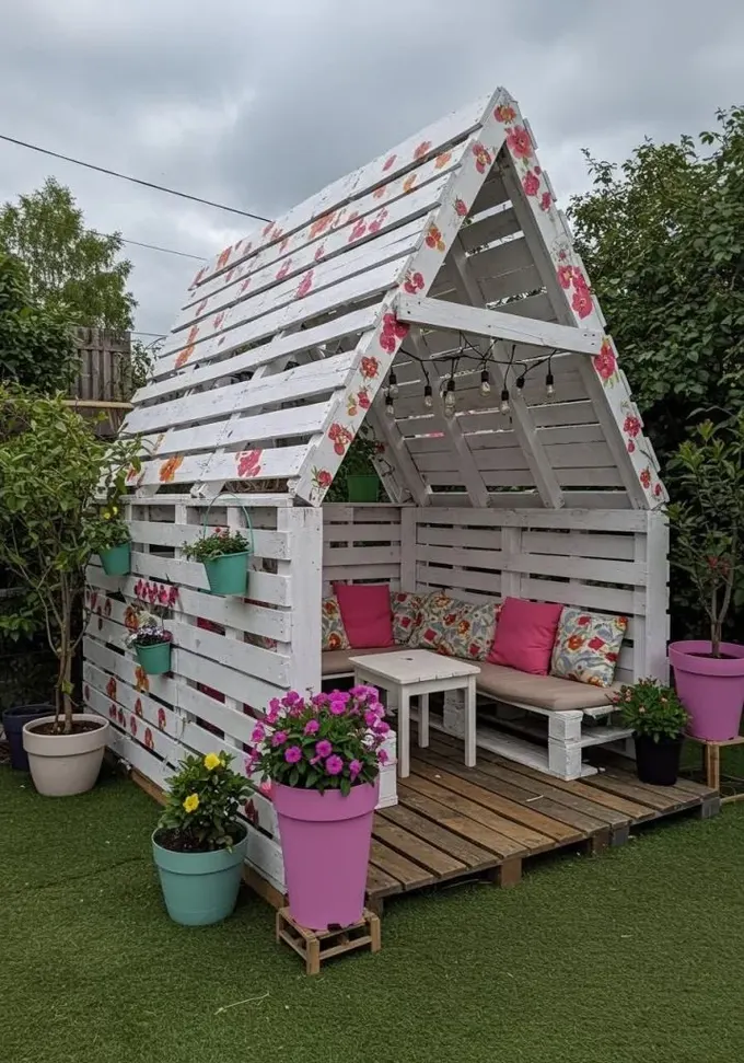 Ev şeklinde palet pergola, üstünde ışıklar ve içinde paletten yapılmış kanepe, canlı çiçekler ve boyalı çiçeklerle tatlı bir alan.
