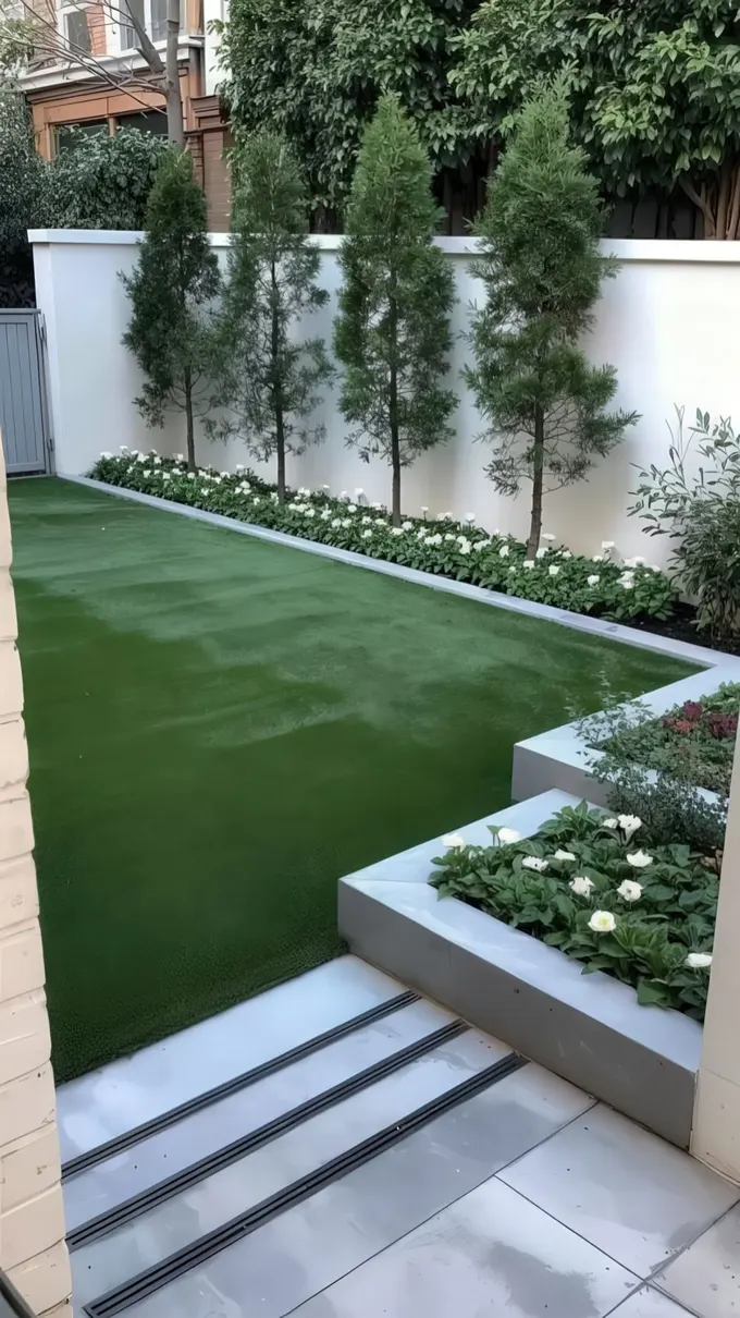 Modern alanda çim ve çit boyunca dar bir bahçe yatağı yer alıyor. Beyaz çiçekler ve uzun ağaçlar çiti şık gösteriyor.