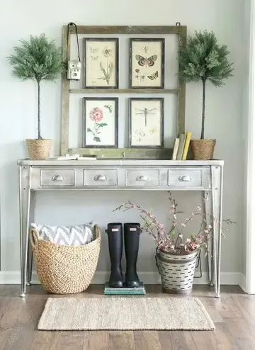 Beyaz boyalı metal konsol, vintage biyoloji posterlerinden oluşan galeri duvarı, saksı bitkileri, kitaplar, lastik botlar ve yapay kiraz çiçeği kova içinde.