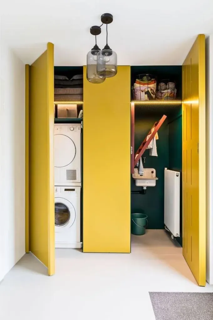 Hardal renkli kapaklı gizli çamaşır odasında bir bölümde makineler, diğer bölümde depolama ve lavabo var.