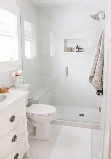 Beyaz mermer karolar, duş, beyaz lavabo ve nötr tonlarda tekstillerle sakin küçük banyo.