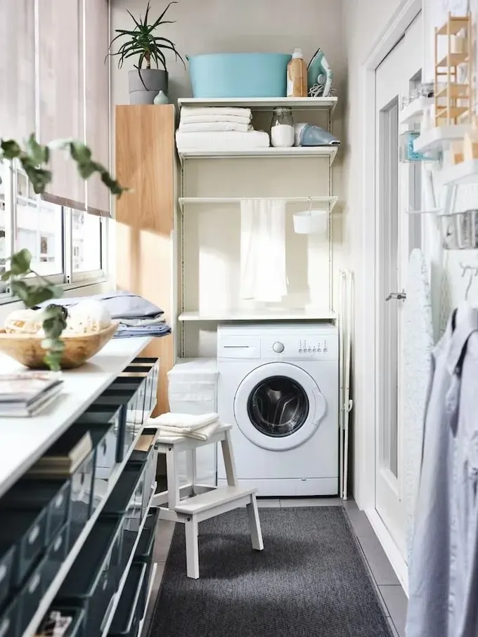 Küçük şehir balkonunda, uzun bir IKEA dolabının içine yerleştirilmiş çamaşır makinesi, hem korunuyor hem de depolama alanı artıyor.