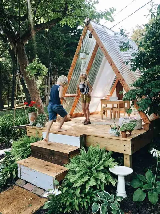 Palet ve camdan yapılmış çocuk evi, küçük mobilya takımı ve çevresinde bol yeşilliklerle tamamlanmış.