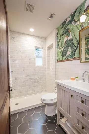 Serin küçük banyo; beyaz mermer ve siyah altıgen karolar, soluk pembe lavabo, tropikal desenli duvar kağıdı ve altın çerçeveli ayna.