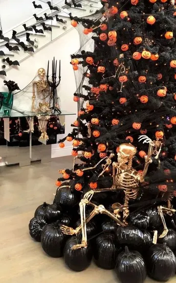 Halloween temalı cesur ve şık bir alan; balkabağı ışıklı ağaç, siyah balkabakları ve altın kaplama iskeletle eğlenceli bir dekorasyon.