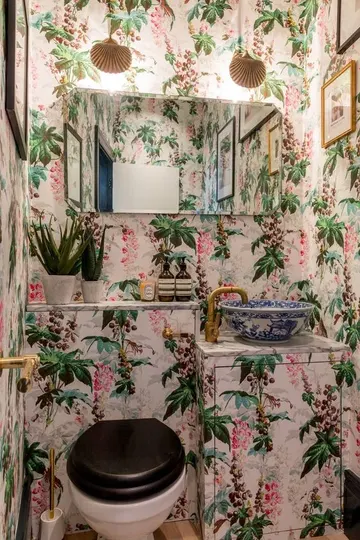 Canlı çiçekli duvar kağıdı, üzerine kaplı lavabo dolabı, ayna, lambalar ve altın detaylarla hareketli misafir tuvaleti.