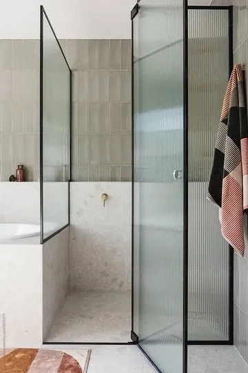 Gri ince ve beyaz taş karolarla huzurlu ve davetkar banyo, siyah çerçeveli oluklu cam kapılar ve tek tokmaklı kapı.