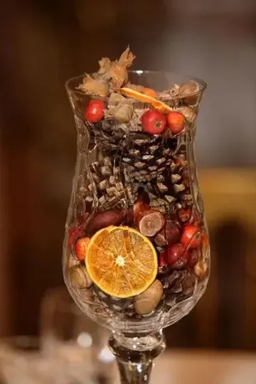 Sahte meyveler, kuruyemişler, kozalaklar ve narenciye dilimleriyle dolu yüksek cam, şık sonbahar dekoru.