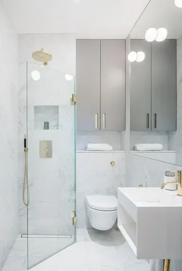 Beyaz mermer karolar, gri dolaplar, altın detaylar ve üçgen duş alanı olan minimal küçük banyo.